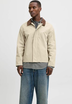 Chaqueta de utilidad beige con un collar marrón, con cremallera frontal y bolsillos laterales, llevada sobre una camisa de cuadros y jeans de pierna ancha.