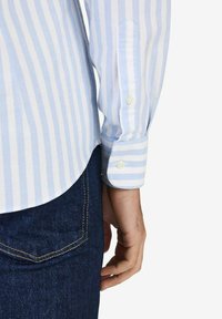 Vaaleansininen ja valkoinen raidallinen pitkähihainen paita napitettavilla hihansuilla, yhdistettynä tummiin farkkuihin. Sileä kangas, joka istuu hyvin.