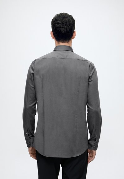 Homme de dos portant une chemise grise à manches longues boutonnée et un pantalon noir sur un fond blanc uni.