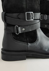 Stivaletti neri in pelle e suede con cinturino in metallo argentato, texture liscia e suola in gomma robusta con dettagli di cucitura visibili.