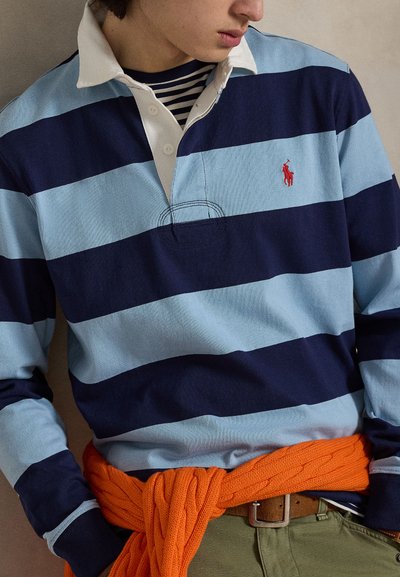 Polo a maniche lunghe a righe azzurro chiaro e blu navy, con colletto bianco e logo rosso. Maglione lavorato a maglia arancione legato in vita.