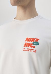 Sudadera blanca con un logo colorido de Nike en rojo y verde azulado, con texto impreso. El tejido parece suave y con una sutil textura. Cuello redondo.