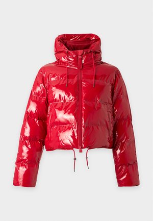 Homme portant une doudoune rouge brillante avec capuche et un pantalon cargo large assorti, chaussures noires, debout sur fond blanc.
