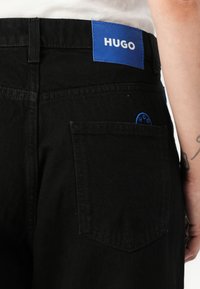 Black denim pants with a blue "HUGO" label at the waistband, visible pocket stitching, and a small circular logo near the back pocket.