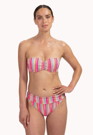 Žena s tmavými vlasy v bezramínkových plavkách s pruhy ve vícero barvách – růžové, oranžové a zelené – pózuje s jednou rukou v bok na bílém pozadí.