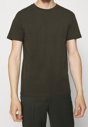 Mand iført en ensfarvet mørkegrøn kortærmet t-shirt og mørke bukser, stående mod en ensfarvet lys baggrund.