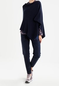 Marin poncho med lös passform, stickad textur, rundad halsringning, lager på ett mönstrat linne. Kombinerad med mörka byxor och grå sportskor.