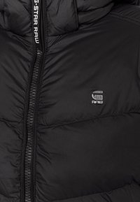Zwarte gewatteerde jas met ritssluiting aan de voorkant en klein zilverkleurig G-STAR RAW logo op de borst nabij de ritssluiting.