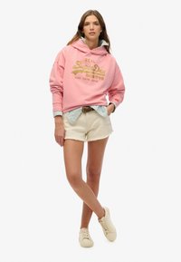 Felpa rosa con logo "SuperDry", indossata sopra una maglietta a righe. Abbinata a pantaloni corti bianchi e sneakers beige. Design semplice e casual.