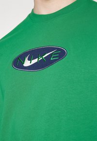 Zelené bavlněné tričko s kulatým výstřihem, s námořnickým oválným nášivkem a vyšitým zeleným logem "NIKE" moderním písmem.