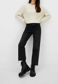 Vrouw in een crèmekleurige ribbeltrui, zwarte hooggeknipte jeans met rafelige zomen en zwarte vetersoldatenlaarzen, staand met handen op de heupen.