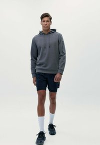 Šedá mikina s kapucňou a šnúrkami, skombinovaná s tmavými šortkami. Model má na sebe čierne tenisky a biele ponožky, stojí pred jednofarebným pozadím.