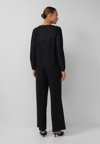 Vrouw met een bun in haar haar, die grote oorbellen draagt, een zwart patroon lange mouwen top, een wijde zwarte broek en zwarte hakken, staande tegen een effen muur.