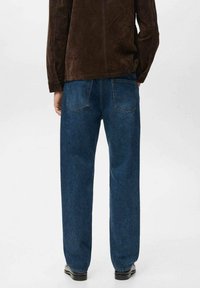 Vaqueros de mezclilla azul oscuro con corte recto, que cuentan con dos bolsillos traseros y un ligero desgaste. Combinados con una chaqueta de ante marrón y zapatos negros.