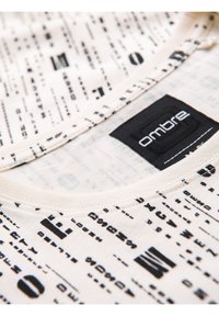 Košile s krátkým rukávem ve světle béžové tkanině s černým abstraktním textovým potiskem. Obsahuje černou logo nášivku na límci.