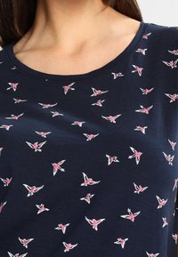 Marinblus med rund halsringning som har ett mönster av små rosa och vita flygande fåglar över tyget.