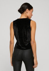 Svart fløyels topp uten ermer med spredte sølvnitter, buet falde og jevn tekstur, kombinert med slanke svarte bukser.