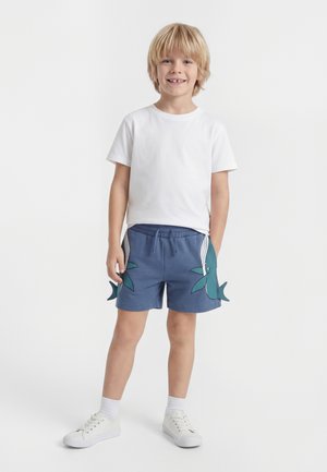 Uśmiechnięty blond chłopiec ubrany w jednolitą białą koszulkę, niebieskie szorty z zielonymi wzorami rekina, białe skarpetki i białe tenisówki, stojący na białym tle.