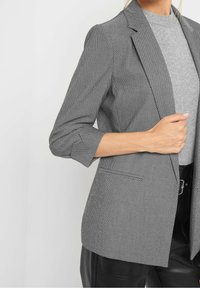 Blazer pied-de-poule noir et blanc avec une coupe ajustée, des revers crantés et des manches trois-quarts, assorti à un haut en maille gris et un pantalon noir.