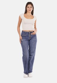 Maglietta aderente di un beige chiaro abbinata a jeans a vita alta blu scuro con chiusura a bottoni e gamba ampia, completata con sneakers bianche.