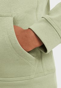Pulover verde deschis cu un finisaj texturat, având un buzunar frontal și manșete cu nervuri, prezentând un braț băgat în buzunar.