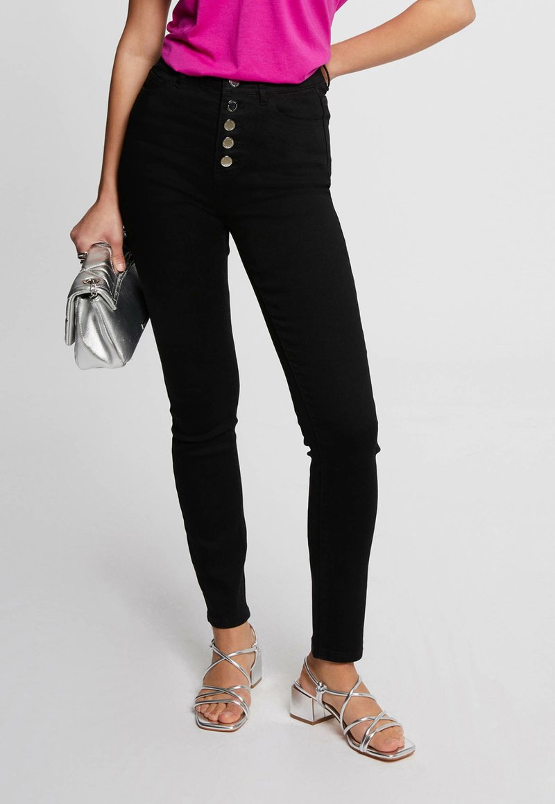 Zwarte high-waisted skinny jeans met een knoopsluiting, aangevuld met zilveren accenten en een glanzende zilveren clutch.