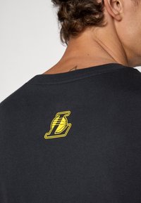 Close-up of a person’s upper back wearing a black shirt with a small yellow basketball and letter "L" logo near the neckline.
