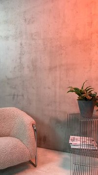 Καμπύλη μπεζ πολυθρόνα με πόδια από χρώμιο δίπλα σε τραπεζάκι με μεταλλικό σκελετό που κρατά γλάστρα και στοίβα από περιοδικά μόδας. Φόντο με τοίχο από σκυρόδεμα.