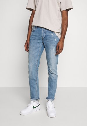 Person iført lyseblå slidte jeans, beige kortærmet skjorte, hvide Nike-sneakers med grønt logo, stående på et ensartet gulv.