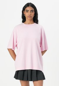 T-shirt oversize rosa chiaro con maniche corte, scollo rotondo e orlo posteriore leggermente più lungo, abbinata a una gonna nera plissettata.