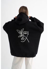 Felpa nera con cappuccio caratterizzata da un disegno a linea bianca di un volto con una foglia. Il tessuto sembra morbido, con una vestibilità ampia e un cappuccio grande.