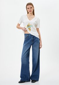 Camiseta blanca de cuello en V con un gráfico dorado y teal en la parte frontal, combinada con unos jeans de mezclilla azul de corte holgado. Unos zapatos negros sólidos completan el conjunto.