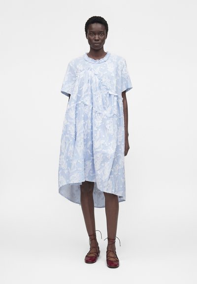 Γυναίκα που φορείται ένα φαρδύ, ανοιχτό μπλε φόρεμα με λευκά ανθισμένα σχέδια και βολάν, σε συνδυασμό με σκούρα κόκκινα, δερμάτινα, επίπεδα δερμάτινα παπούτσια με κορδόνια, στέκεται μπροστά σε ένα μονόχρωμο φόντο.