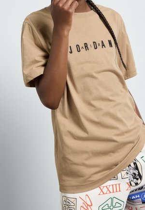 Persona che indossa una maglietta Jordan beige con scritte nere e pantaloni bianchi con motivi grafici multicolori e numeri.