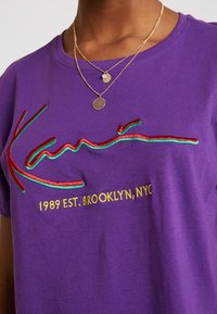 Lila bomull t-shirt med röd och grön broderi av "Kan's" logotyp och texten "1989 EST. BROOKLYN, NYC" på framsidan.
