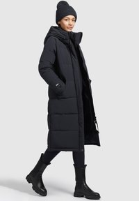 Abrigo acolchado negro con capucha, hasta la rodilla, con un diseño de acolchado y bolsillos laterales, combinado con botas negras hasta el tobillo y un gorro de canalé.