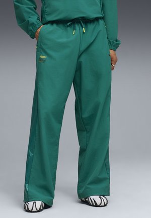 Person iført løse grønne træningsbukser med gule snore og logo på lommen, kombineret med hvide og sorte sneakers.