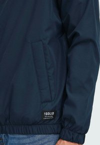 Navy blue lightweight jacket with a smooth texture, elastic cuffs, side pocket, and a branded tag at the hem displaying the slogan.
