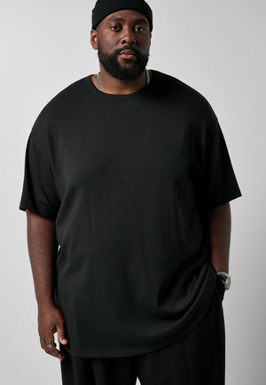 Mężczyzna z brodą, ubrany w czarną czapkę, czarny oversize t-shirt, srebrny łańcuszek, zegarek na rękę i pierścionek, stoi na tle prostego, szarego tła.