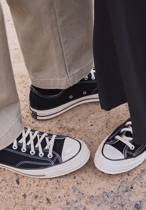 Schwarze Segeltuch-Sneaker mit weißen Gummi-Zehenkappen und Schnürsenkeln. Kontrastnähte und Metallösen geben dem Schuh Detail. Getragen mit beigen Hosen und einem schwarzen Stoff.