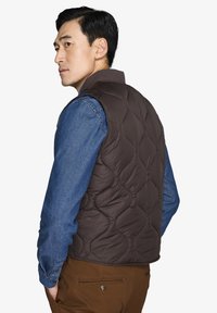 Gilet imbottito marrone con colletto alto, indossato sopra una camicia in denim. Il gilet presenta una texture liscia e un design aderente.