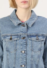 Chaqueta de mezclilla azul clara con un collar clásico, botones plateados y dos bolsillos en el pecho con costuras en contraste. Textura suave, ajuste entallado.