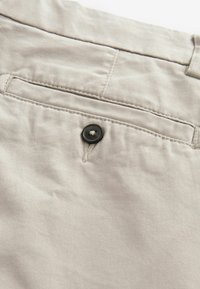 Tissu beige à texture lisse, doté d'une poche arrière et d'un détail bouton noir. Les coutures sont visibles le long des bords.