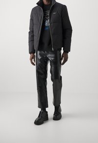 Sort pufferjakke med høj krave og lynlås, båret over en sort sweatshirt med en blå grafik. Sorte læderbukser og sko fuldender outfittet.