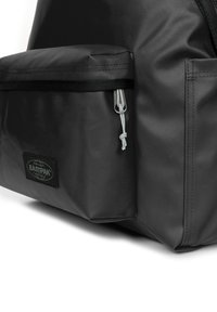 Mochila de tela negra con un bolsillo frontal, cierre con cremallera y cordón gris. Incluye un parche de logo negro de Eastpak cosido.