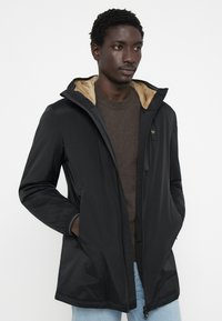 Schwarze, leichte, wasserabweisende Jacke mit einem weichen, tanfarbenen Fleece-Innenfutter, verstellbarer Kapuze und zwei seitlichen Taschen. Reißverschluss vorne.