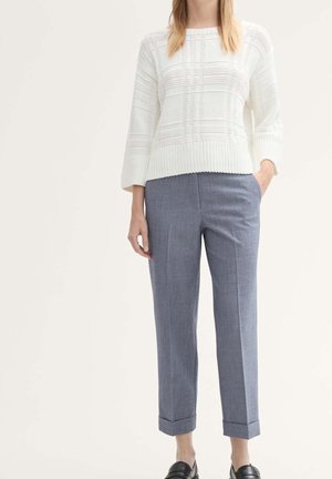 Mujer con un jersey de punto blanco con textura, pantalones grises a medida hasta el tobillo y zapatos negros sin cordones, de pie con una mano en el bolsillo.