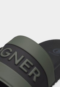 Κοντινό πλάνο σε λουρί μαύρης και ελιάς πράσινης σαγιονάρας με ανασηκωμένα γράμματα "GANT" στο λουρί.