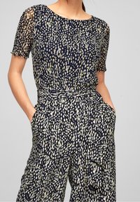 Marinblå jumpsuit med korta ärmar, lös passform och en texturerad tyg med ett ljust prickmönster i kräm och grönt. Elastisk midja med fickor.