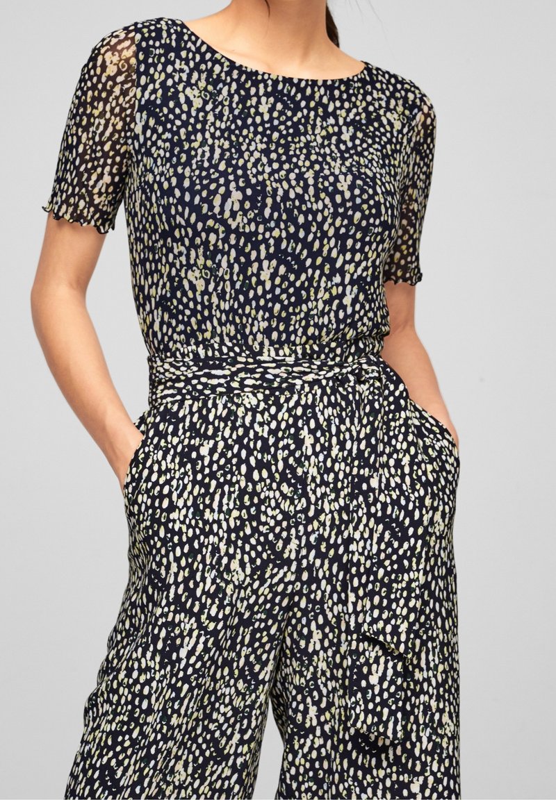 Marinblå jumpsuit med korta ärmar, lös passform och en texturerad tyg med ett ljust prickmönster i kräm och grönt. Elastisk midja med fickor.